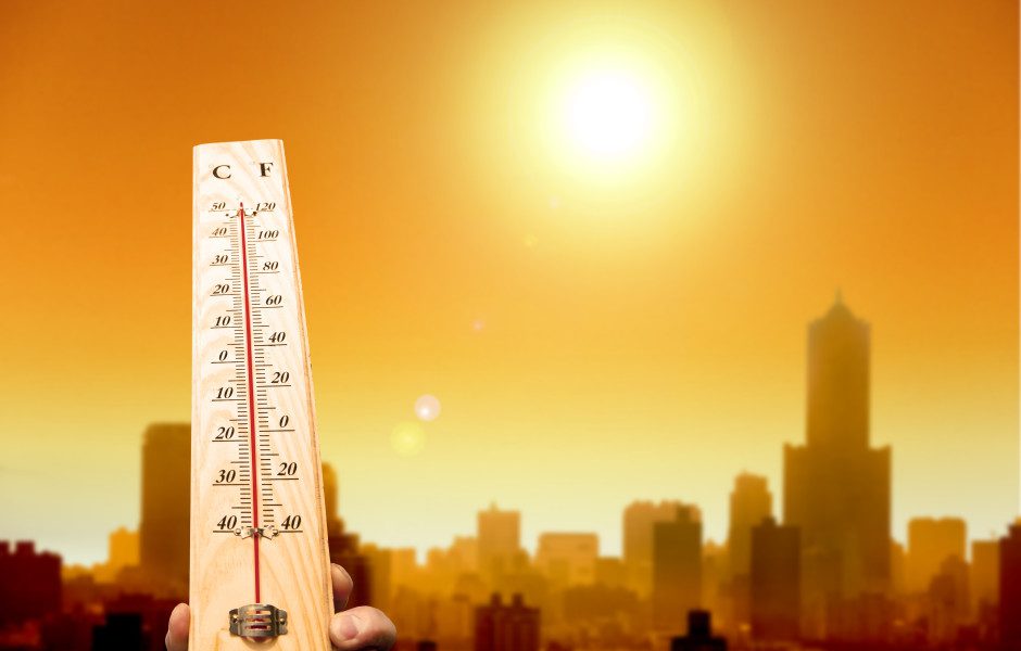 A thermometer in front of the sun with buildings behind it.